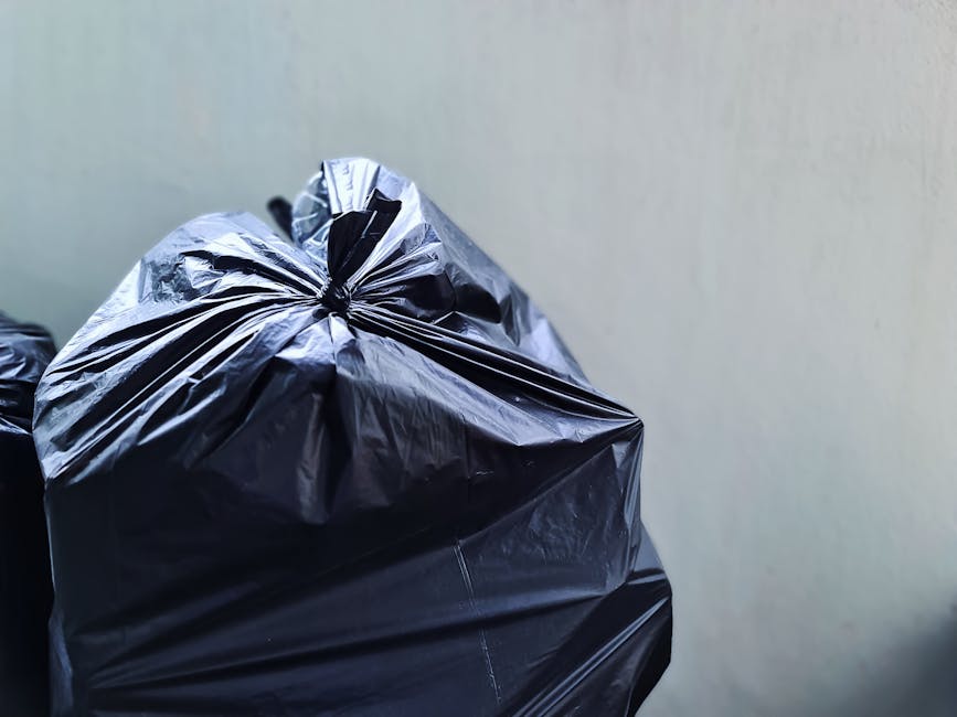 Close-up view of a large black plastic rubbish bag tightly tied at the top, positioned in the foreground against a plain, light-colored wall background. The bag appears to be filled with household was