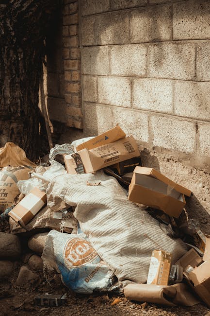 Close-up view of a large black plastic rubbish bag tightly tied at the top, positioned in the foreground against a plain, light-colored wall background. The bag appears to be filled with household was
